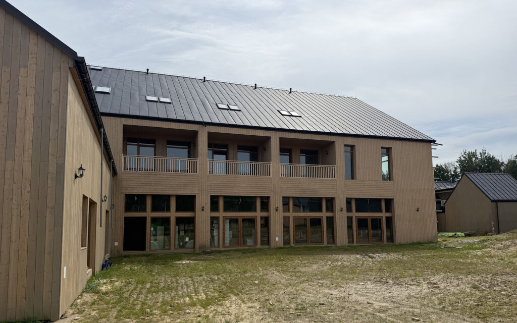 Na zdjęciu znajduje się nowoczesny, dwukondygnacyjny budynek o elewacji z jasnego drewna i dachu pokrytym blachą na rąbek. Duże przeszklenia na parterze oraz zadaszony balkon z drewnianą balustradą na piętrze nadają konstrukcji lekkości i otwartości. Na dachu widoczne są świetliki, które dodatkowo doświetlają wnętrze. Otoczenie to trawiasty teren z elementami ziemi, a w tle widać inne budynki o podobnej stylistyce.