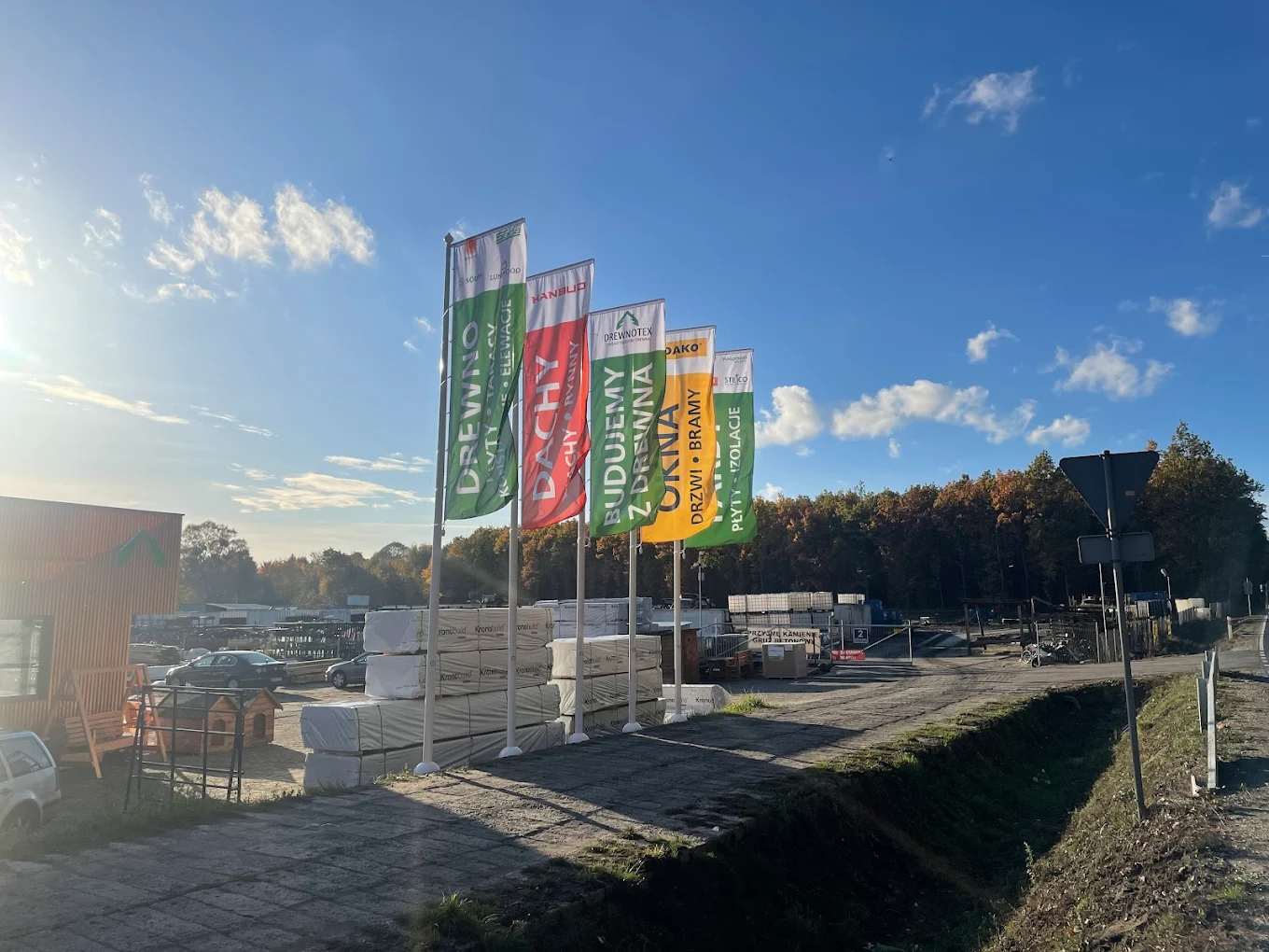 Na zdjęciu widzimy zewnętrzny teren składu budowlanego lub centrum dystrybucji materiałów konstrukcyjnych. W tle powiewają kolorowe flagi reklamowe z polskimi napisami takimi jak „DREWNO”, ,,OKNA'', „BUDUJEMY Z DREWNA” czy „KONSTRUKCJE DACHOWE”, które jednoznacznie wskazują na specjalizację firmy w drewnie konstrukcyjnym i dachowym. Przestrzeń jest zorganizowana — widoczne są stosy drewna, palety i różne materiały budowlane.