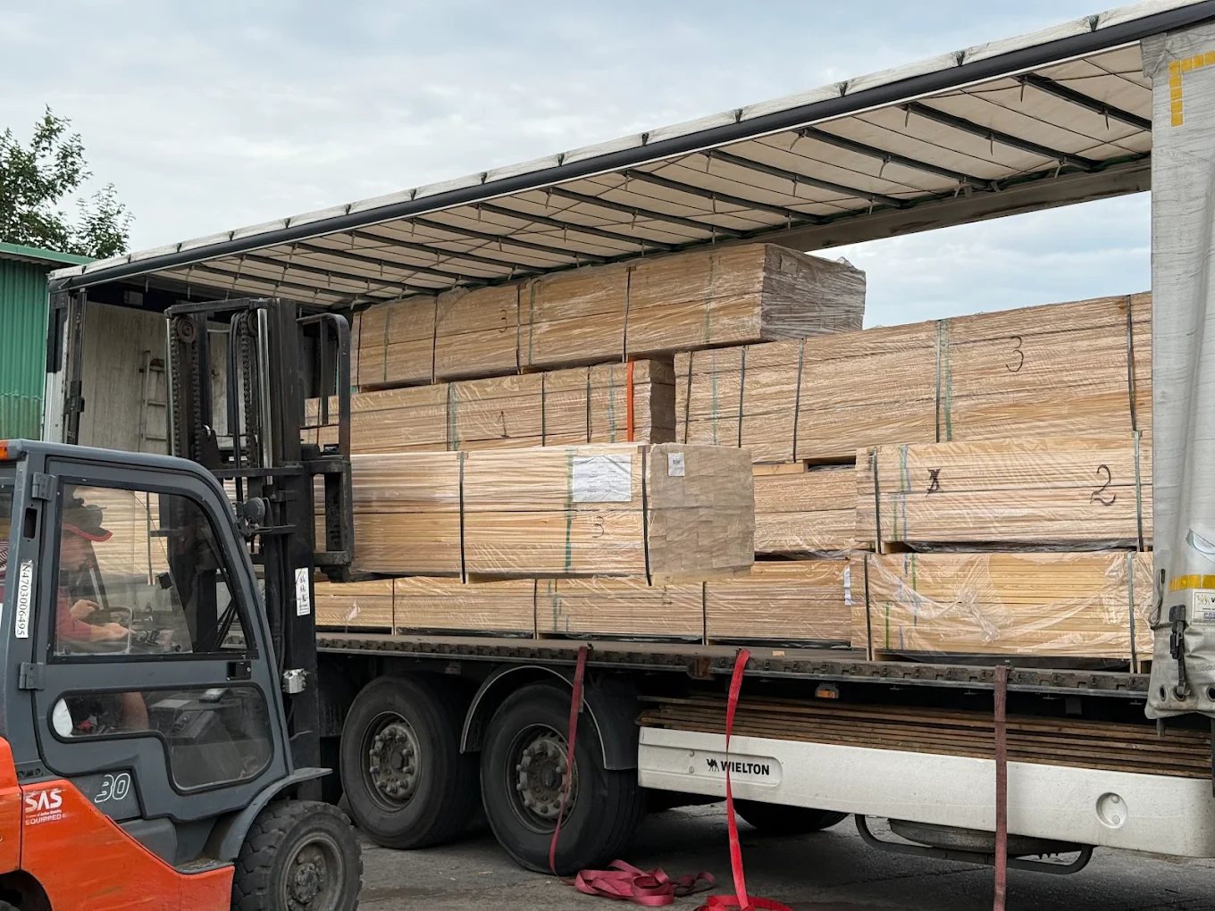 Na zdjęciu widzimy dynamiczną scenę załadunku lub rozładunku materiałów budowlanych. W centrum znajduje się wózek widłowy obsługiwany przez operatora, który manewruje przy otwartej naczepie ciężarówki. Wewnątrz naczepy znajdują się starannie ułożone i owinięte folią pakiety drewnianych płyt, oznaczone etykietami i numerami identyfikacyjnymi. Czerwone pasy zabezpieczające zwisają z boku, świadcząc o dbałości o stabilność ładunku podczas transportu.