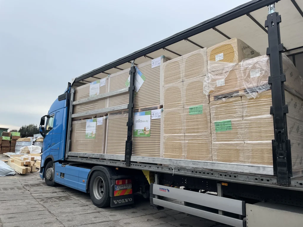 Na zdjęciu widzimy niebieską ciężarówkę marki Volvo z otwartą naczepą, załadowaną dużymi pakietami drewnianych płyt lub paneli. Materiały są starannie ułożone i owinięte folią ochronną, z widocznymi etykietami informacyjnymi. Pojazd stoi na utwardzonym placu, prawdopodobnie przy magazynie lub centrum dystrybucji. Całość ukazuje profesjonalną organizację transportu materiałów budowlanych.